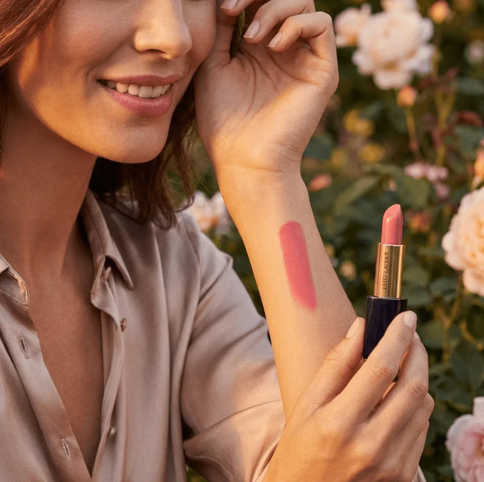 Woman showing pink lipstick swatch on arm, holding lipstick outdoors among flowers