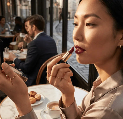 Woman applying dark red matte lipstick in a cafe setting with coffee and pastries on table