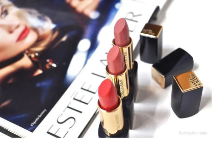 Three pink and red matte lipsticks with gold and black tubes, shown on a magazine background.