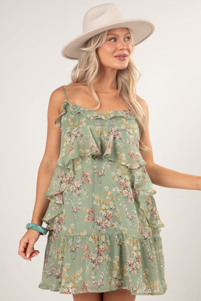 A woman wearing a sage green tiered ruffle mini dress with a pink and yellow floral print, styled with a beige wide-brim hat and a turquoise bracelet against a light gray studio background.