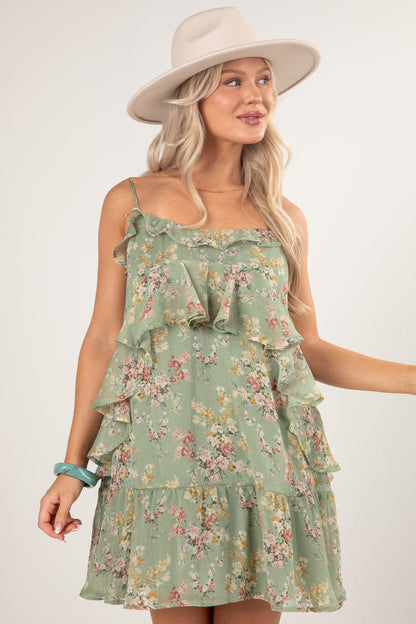 A woman wearing a sage green tiered ruffle mini dress with a pink and yellow floral print, styled with a beige wide-brim hat and a turquoise bracelet against a light gray studio background.