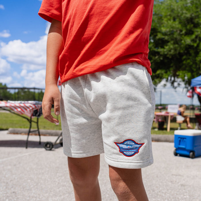 Lower body of youth male model wearing heather gray Fastball Flight sweatshorts with red tee showing Air Show Division badge on leg at outdoor baseball event