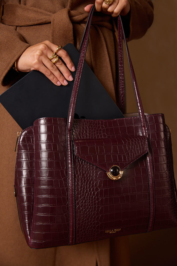 Female model holding the Francois Black Cherry Crocodile print faux leather Laptop Tote by the shoulder straps and putting a laptop into the central laptop zipper compartment