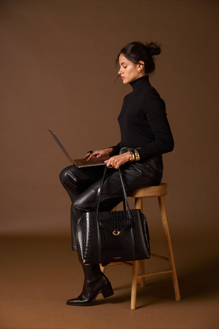 Female model sitting with her laptop on her lap and holding Francois Black Crocodile print faux leather Laptop Tote by her side using the two top shoulder straps