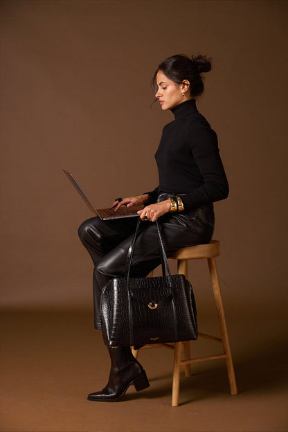 Female model sitting with her laptop on her lap and holding Francois Black Crocodile print faux leather Laptop Tote by her side using the two top shoulder straps