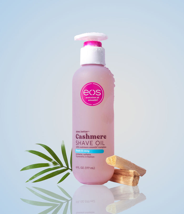 Pink bottle of eos Cashmere Shave Oil with a palm leaf and wooden applicator on a light blue background