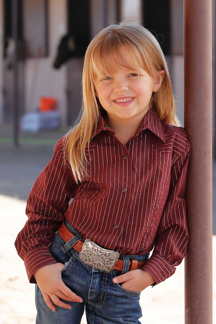 Young girl leaning on a metal pole wearing a Cruel Denim Burgundy with gold metallic stripe snap Long Sleeve Western Shirt with jeans and a belt. Front view.