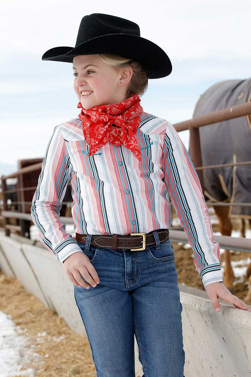 A young girl is wearing a turquoise/red/ black striped Cinch long sleeve shirt. She is also wearing a black cowboy hat, a red printed wild rag, jeans, and a belt. Front view of her standing beside an outdoor feed trough.