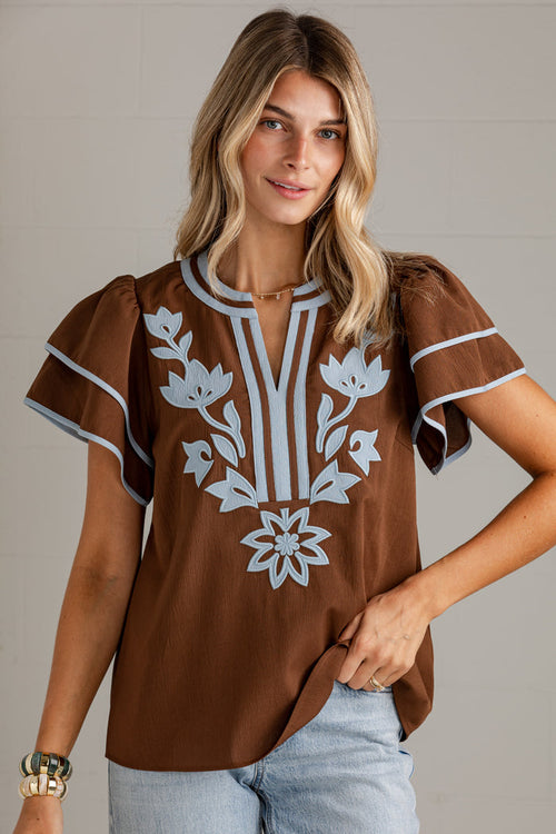 A woman wearing a brown short-sleeve blouse with white floral embroidery and striped trim, paired with light blue jeans, against a neutral gray background.