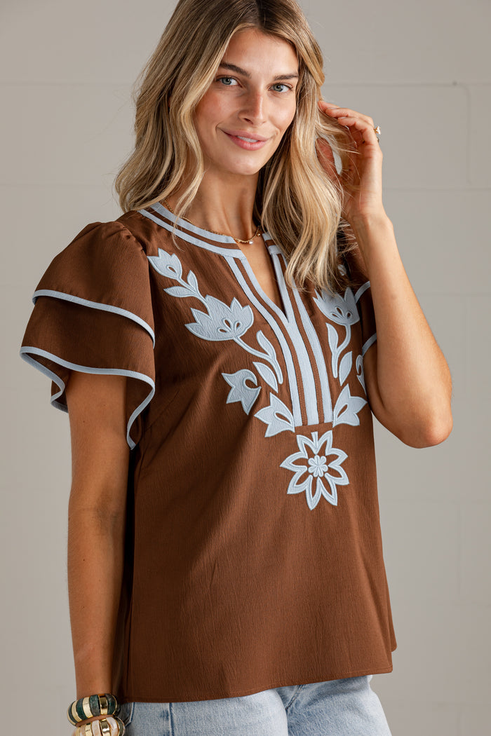 A woman wearing a brown short-sleeve top with white floral embroidery on the front, paired with light blue jeans, photographed against a neutral light gray background.