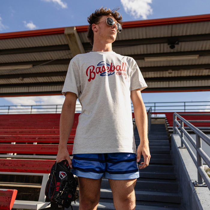Man holding a glove at red stadium bleachers looking up wearing aviator sunglasses a white heather Baseball Lifestyle graphic tee and royal blue striped mesh shorts