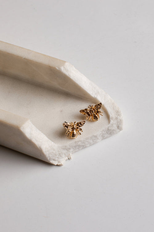 A pair of small gold bee stud earrings resting on a white ceramic tray against a light gray background.
