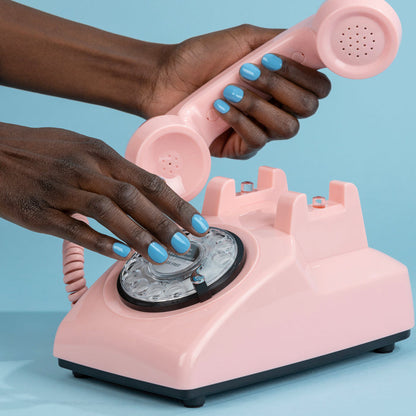 Close-up of hands with Hot Spring Pacific blue-painted nails dialing a vintage pink rotary phone against a light, pool-blue background, brought to you by LONDONTOWN.