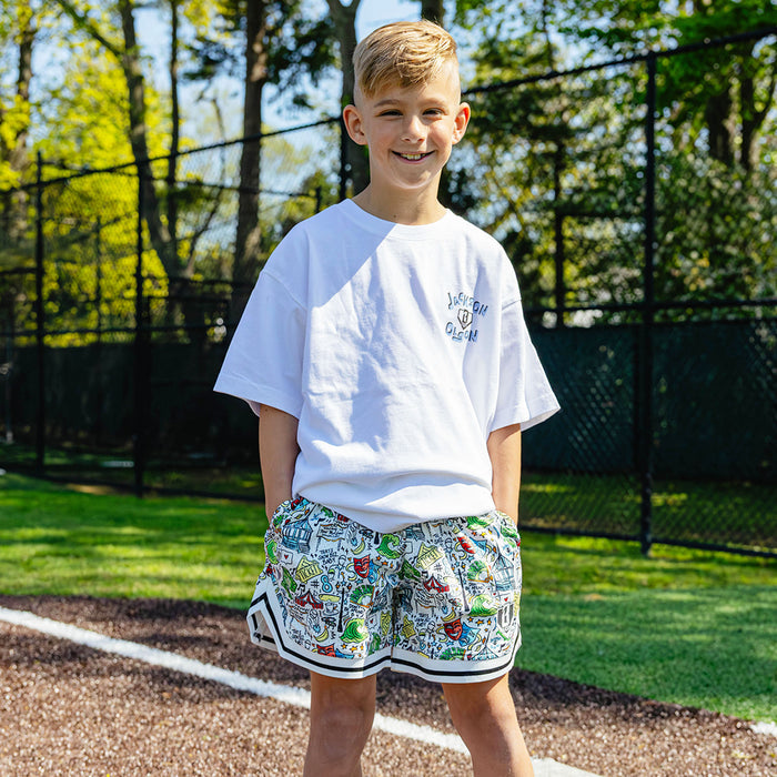 Smiling youth player on a baseball field wearing the white Jackson Olson x BL101 Hometown Shorts with all-over illustrated print paired with the white Jackson Olson Hometown Relaxed Tee