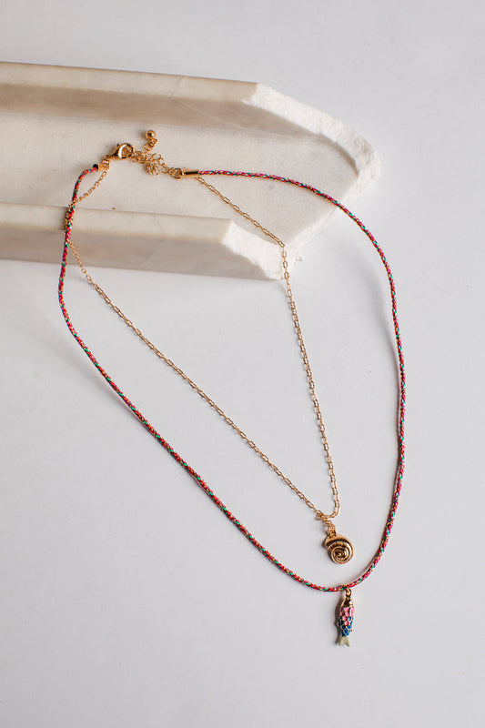 A delicate layered necklace set featuring a gold chain with a small gold spiral pendant and a longer red-and-gold beaded chain with a small multicolor fish charm, displayed on a light gray surface next to a white ceramic tray.
