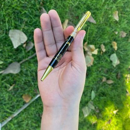 Hand holding a black and gold pen against a grassy background