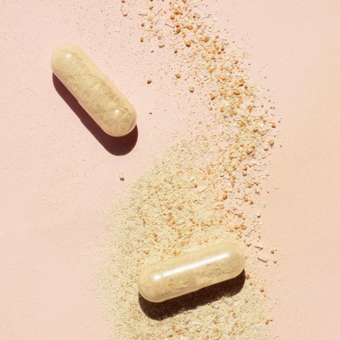 Two beige Nail Boost capsules by Manucurist sit on a light pink surface, partly covering scattered beige powder—ideal for those seeking supplements to strengthen nails and support healthy nail growth.