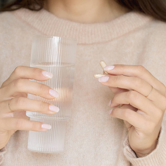 A person in a light pink sweater holds a ribbed glass of water and two Manucurist Nail Boost supplements, preparing to take the medication to help strengthen their nails.