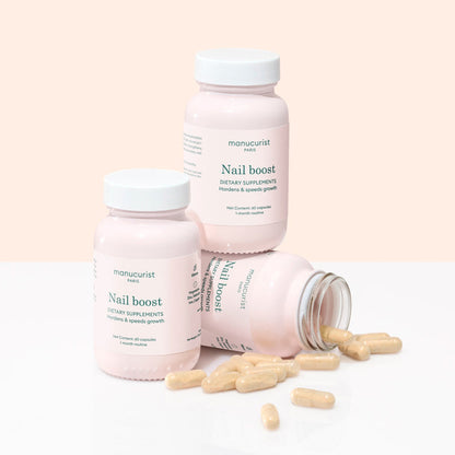 Three bottles of Manucurist Nail Boost supplements are arranged on a light surface, one lying on its side with beige capsules spilled out, highlighting support for nail growth and stronger nails.