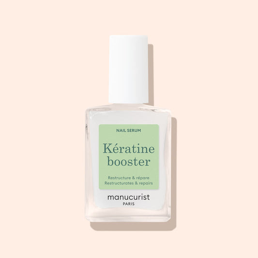 A clear glass bottle of Manucurist Nail Strengthening Serum with a white cap and green label, highlighting its strengthening benefits for damaged nails, is displayed against a pale beige background.