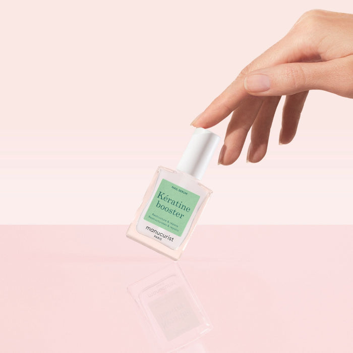 A manicured hand balances a bottle of Manucurist Nail Strengthening Serum on a glossy pink surface with a soft pink background—ideal for reviving and caring for damaged nails.