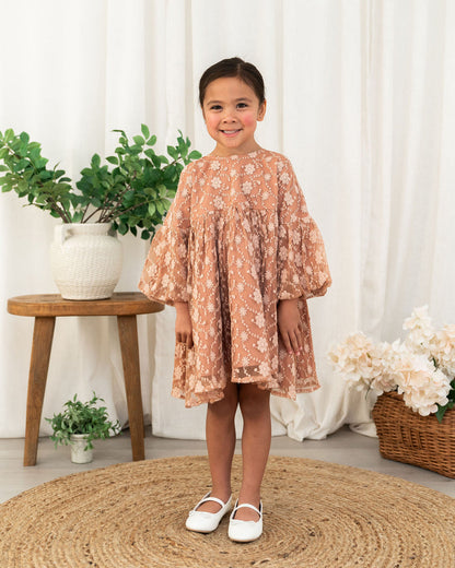 A toddler girl stands smiling in a dusty rose organic lace dress with white floral embroidery, paired with white Mary Jane shoes, set against a soft white curtain backdrop with decorative plants and a woven rug.