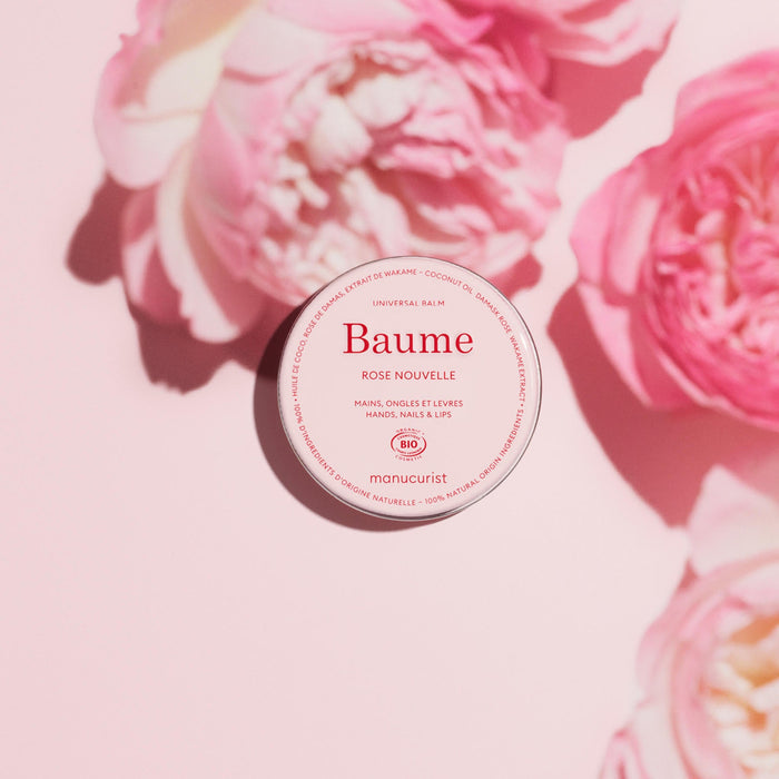 A round tin of Manucurist Natural Nourishing Balm - Cosmos Organic sits on a light pink surface, surrounded by soft pink flowers, creating a delicate and feminine look.