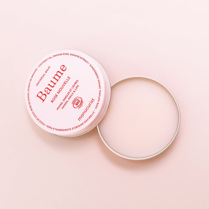 A round, open tin of Manucurist Natural Nourishing Balm - Cosmos Organic rests on a light pink surface. The lid with red branding lies beside it, showing the smooth balm inside—perfect for lip repair and natural skincare.