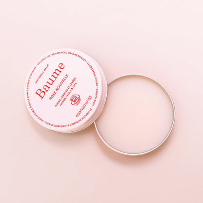 A round, open tin of Manucurist Natural Nourishing Balm - Cosmos Organic rests on a light pink surface. The lid with red branding lies beside it, showing the smooth balm inside—perfect for lip repair and natural skincare.