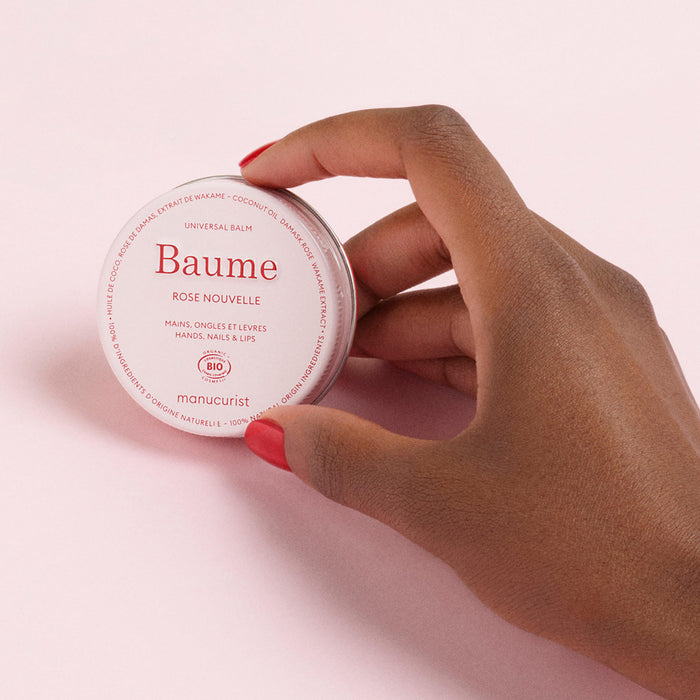 A hand with red-painted nails holds a round tin of Manucurist Natural Nourishing Balm - Cosmos Organic against a light pink background. This balm nourishes hands, nails, and lips, making it ideal for natural skincare and lip repair.