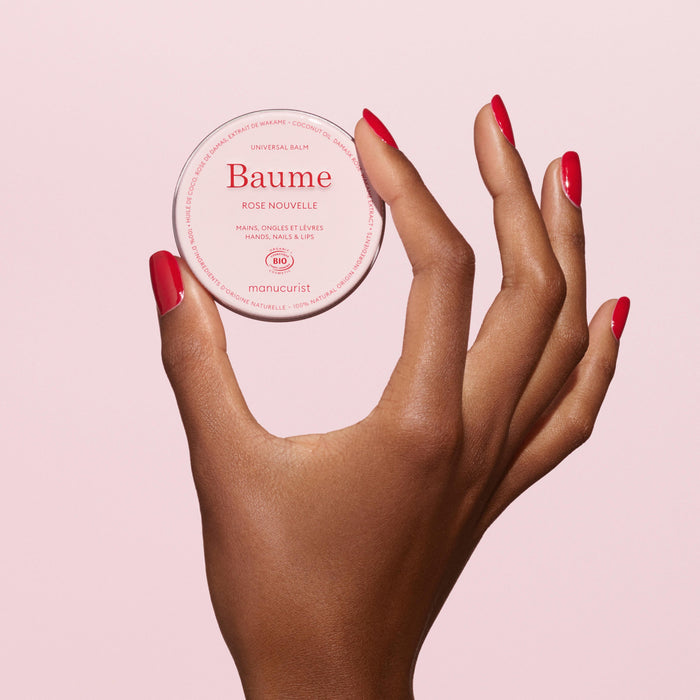 A hand with neatly manicured, red nails holds up a round container of Manucurist Natural Nourishing Balm - Cosmos Organic for natural skincare and cuticle care against a light pink background.