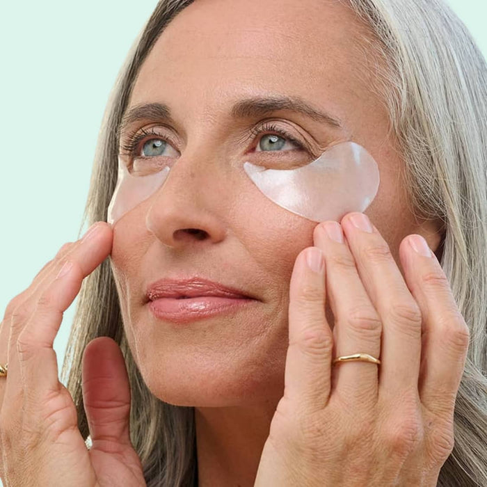 Woman with long silver hair wearing clear under-eye gel patches, pressing fingertips to her cheeks against a mint background