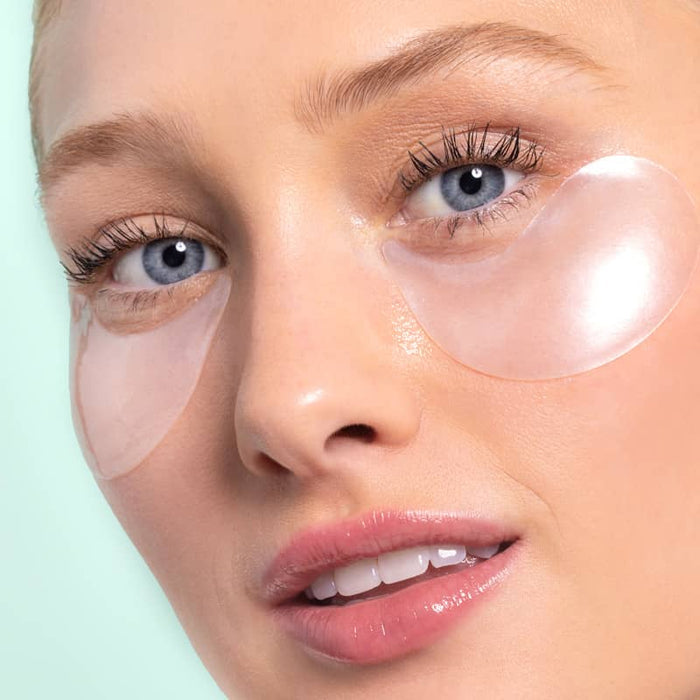 Close-up of a woman with blue eyes and glossy pink lips wearing clear hydrogel under-eye patches against a mint background