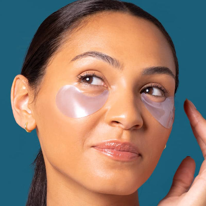 Woman with dark slicked-back hair wearing translucent under-eye gel patches, glancing upward against a dark teal background