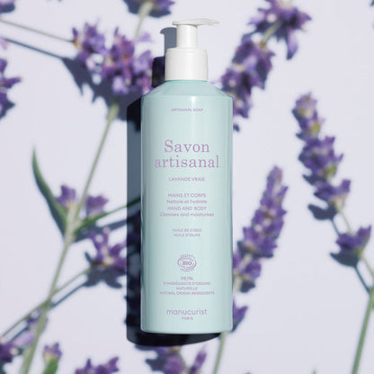 A light blue bottle of Manucurist Organic Lavender Hand  Body Wash - Cosmos Organic with a pump sits on a surface with lavender flowers and soft shadows. The label highlights coconut and olive oil, natural ingredients, and a subtle lavender scent.