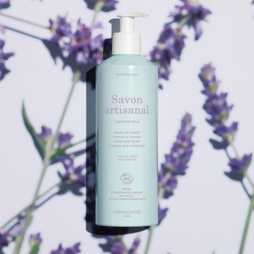 A light blue bottle of Manucurist Organic Lavender Hand  Body Wash - Cosmos Organic with a pump sits on a surface with lavender flowers and soft shadows. The label highlights coconut and olive oil, natural ingredients, and a subtle lavender scent.
