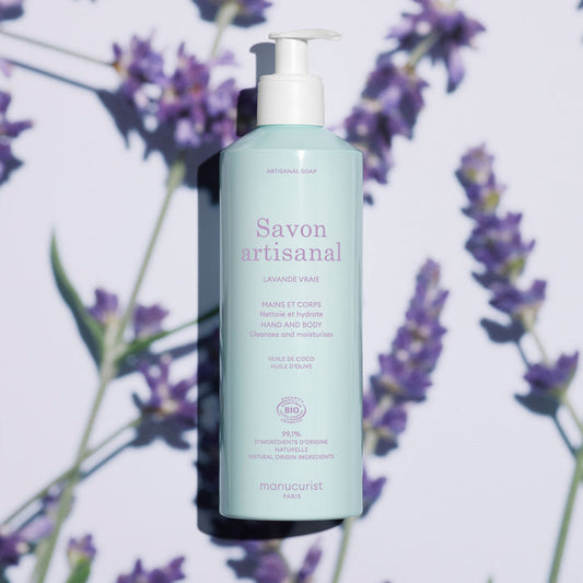 A light blue bottle of Manucurist Organic Lavender Hand  Body Wash - Cosmos Organic with a pump sits on a surface with lavender flowers and soft shadows. The label highlights coconut and olive oil, natural ingredients, and a subtle lavender scent.