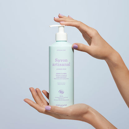 A person with lavender-painted nails holds a light blue pump bottle of Manucurist’s Organic Lavender Hand  Body Wash - Cosmos Organic against a light blue background, suggesting a soothing lavender scent.