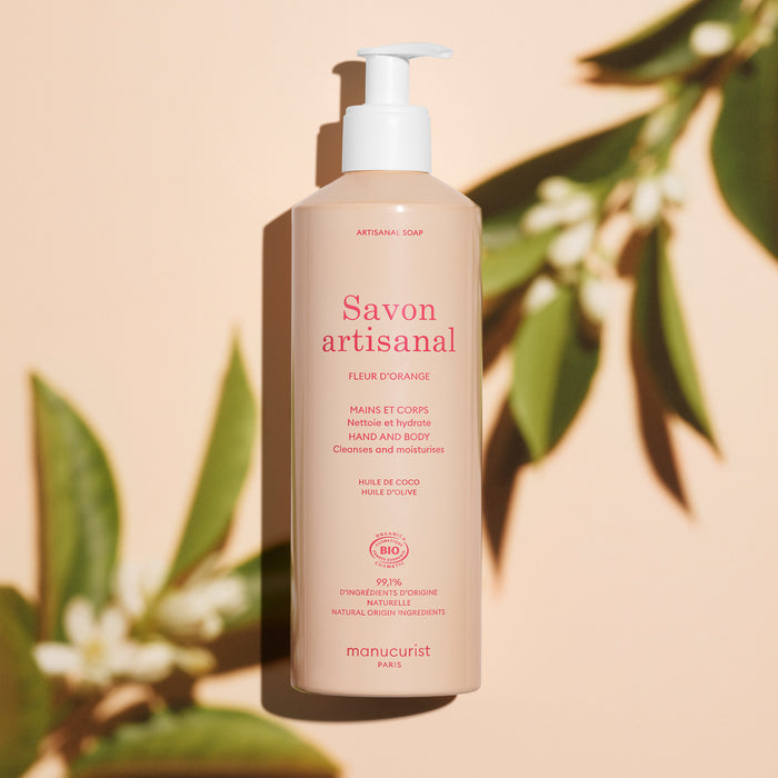 A beige pump bottle of Manucurist Organic Orange Blossom Hand  Body Wash - Cosmos Organic stands upright on a peach background, surrounded by green leaves and their shadows, showcasing this artisan soap infused with orange blossom.