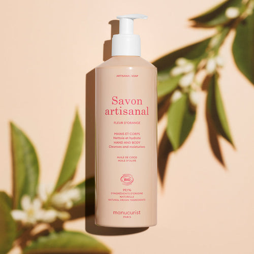 A beige pump bottle of Manucurist Organic Orange Blossom Hand  Body Wash - Cosmos Organic stands upright on a peach background, surrounded by green leaves and their shadows, showcasing this artisan soap infused with orange blossom.