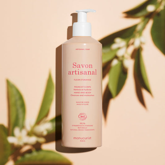 A beige pump bottle of Manucurist Organic Orange Blossom Hand  Body Wash - Cosmos Organic stands upright on a peach background, surrounded by green leaves and their shadows, showcasing this artisan soap infused with orange blossom.