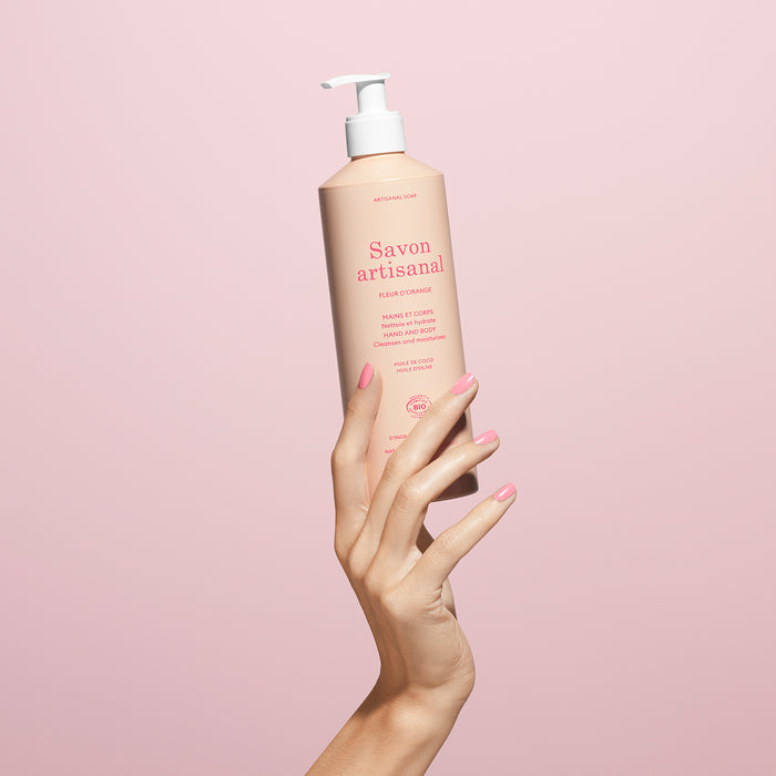 A manicured hand holds a pink bottle of Manucurist Organic Orange Blossom Hand  Body Wash – Cosmos Organic, set against a pale pink background.