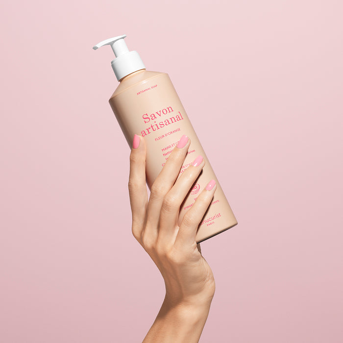 A hand with manicured pink nails holds up a bottle of Manucurist Organic Orange Blossom Hand  Body Wash - Cosmos Organic with a pump dispenser against a pale pink background.