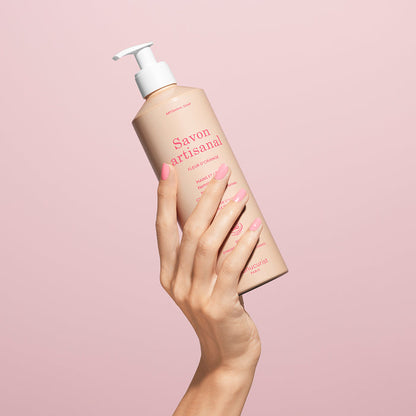 A hand with manicured pink nails holds up a bottle of Manucurist Organic Orange Blossom Hand  Body Wash - Cosmos Organic with a pump dispenser against a pale pink background.