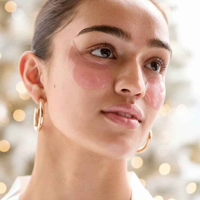 Close-up of a woman wearing pink gel under-eye patches and gold hoop earrings against soft holiday lights