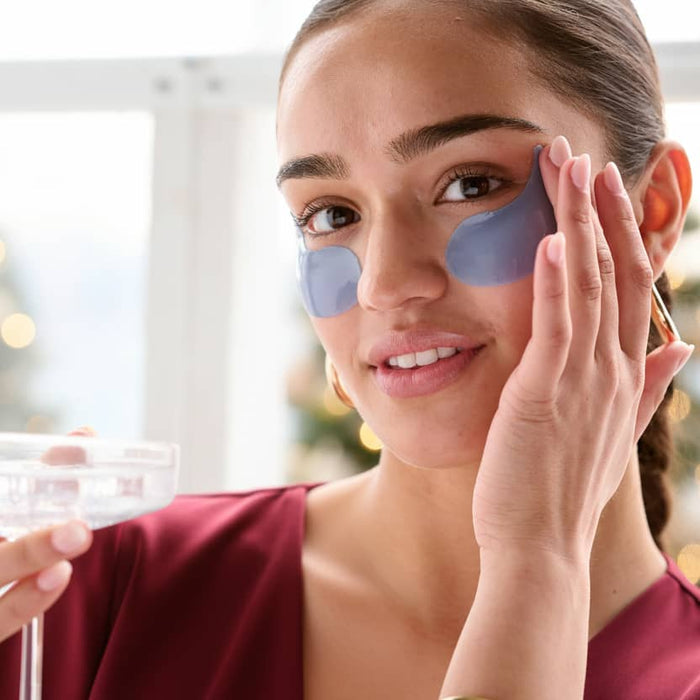 Smiling woman in a burgundy top pressing blue gel eye patches under her eyes while holding a coupe glass near a holiday tree
