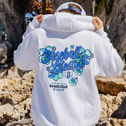 Person wearing a white hoodie with floral 'Baseball Lifestyle Swing into Spring Beach Club' design outdoors.
