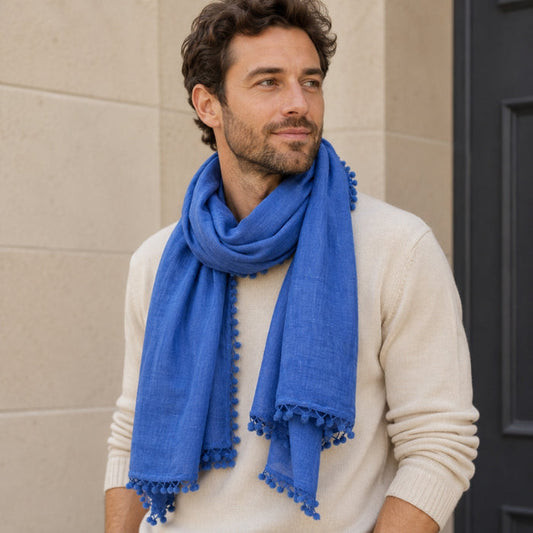 Man wearing a blue scarf with pom-pom details, standing against a beige wall.