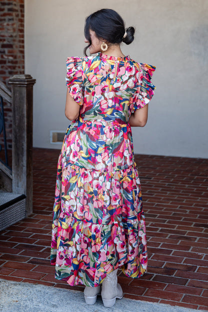 A woman viewed from behind wearing a colorful floral midi dress with ruffled short sleeves and a tiered skirt, standing on a brick patio against a stucco wall.