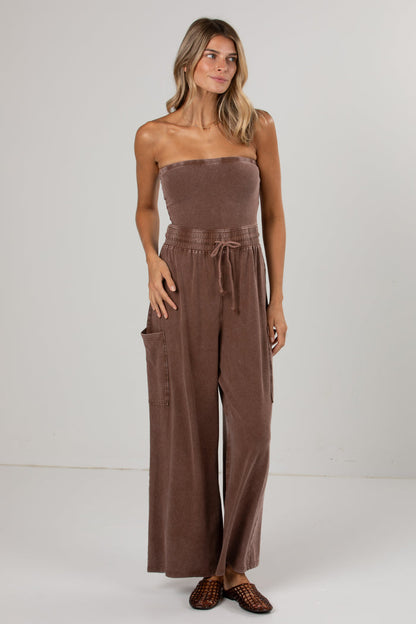 A woman modeling a brown strapless wide-leg jumpsuit with a drawstring waist and side pockets, standing against a plain light gray studio background.
